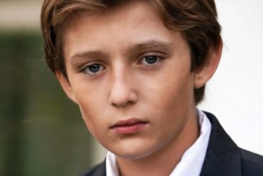 Young man in formal black suit and white shirt, portrait photograph related to Barron Trump upbringing privacy discussion