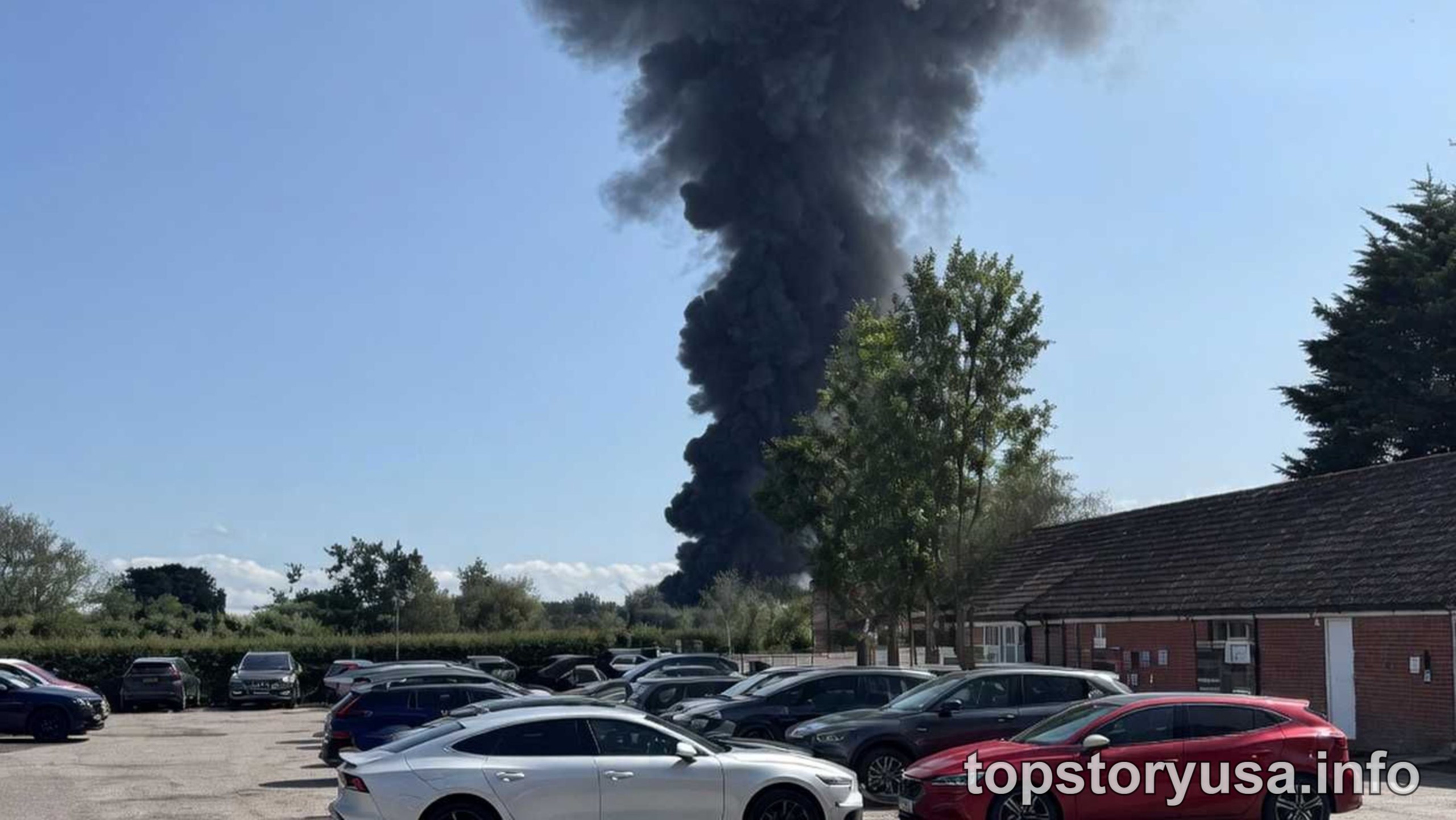 BREAKING NEWS large dark smoke plume rising from industrial fire in parking lot area with vehicles and buildings